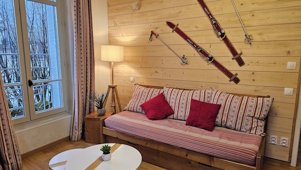 A cozy room with a wooden wall, a sofa with patterned cushions, a small table with a plant, and a window with curtains.