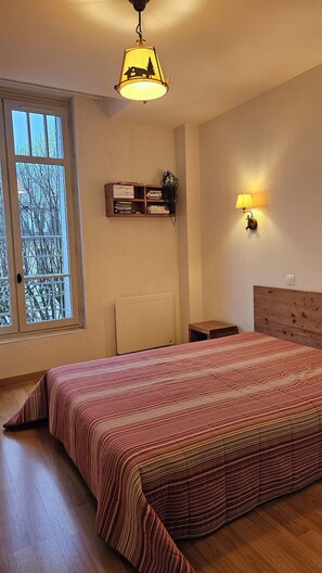 A bedroom with a bed, a window, a wall-mounted shelf with books, a bedside lamp, and a ceiling light.