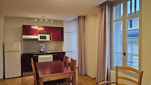 A compact kitchen with a microwave, refrigerator, and coffee maker. There is a dining table with chairs and a window with curtains.