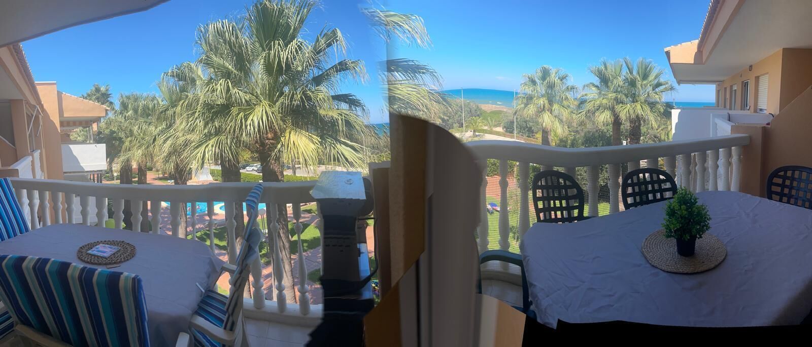 Ein Balkon mit Blick auf Palmen und das Meer. Zwei Tische mit Stühlen stehen bereit, einer mit einem Streifen-Kissen und der andere mit einer weißen Tischdecke und einer kleinen Pflanze.