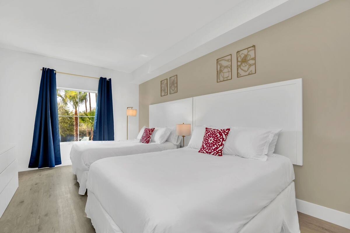 A hotel room with two beds, a large window with curtains, a beige wall with framed artwork, and a wooden floor.