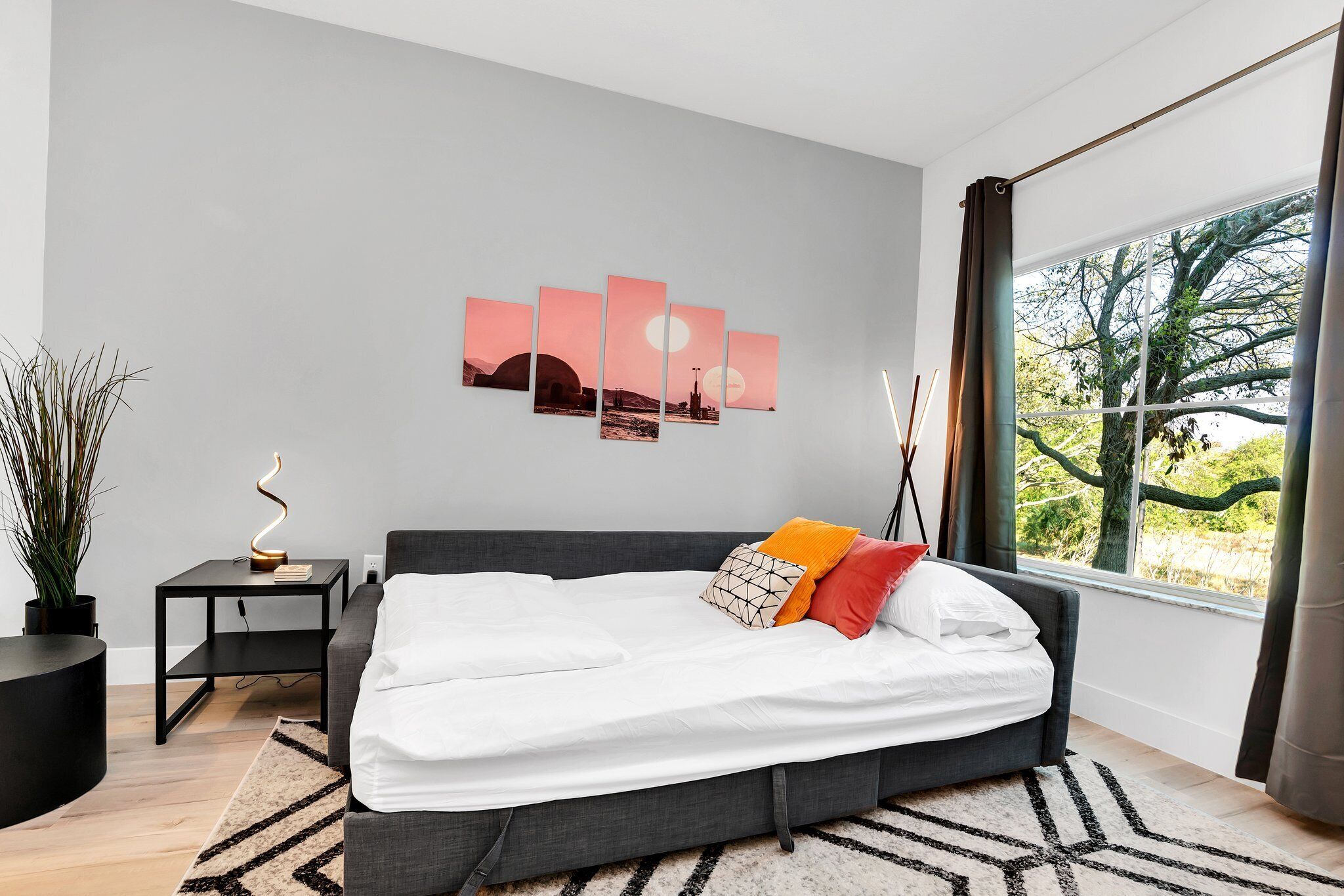 A modern bedroom with a large bed, a bedside table, a wall-mounted artwork, and a view of trees through a window.