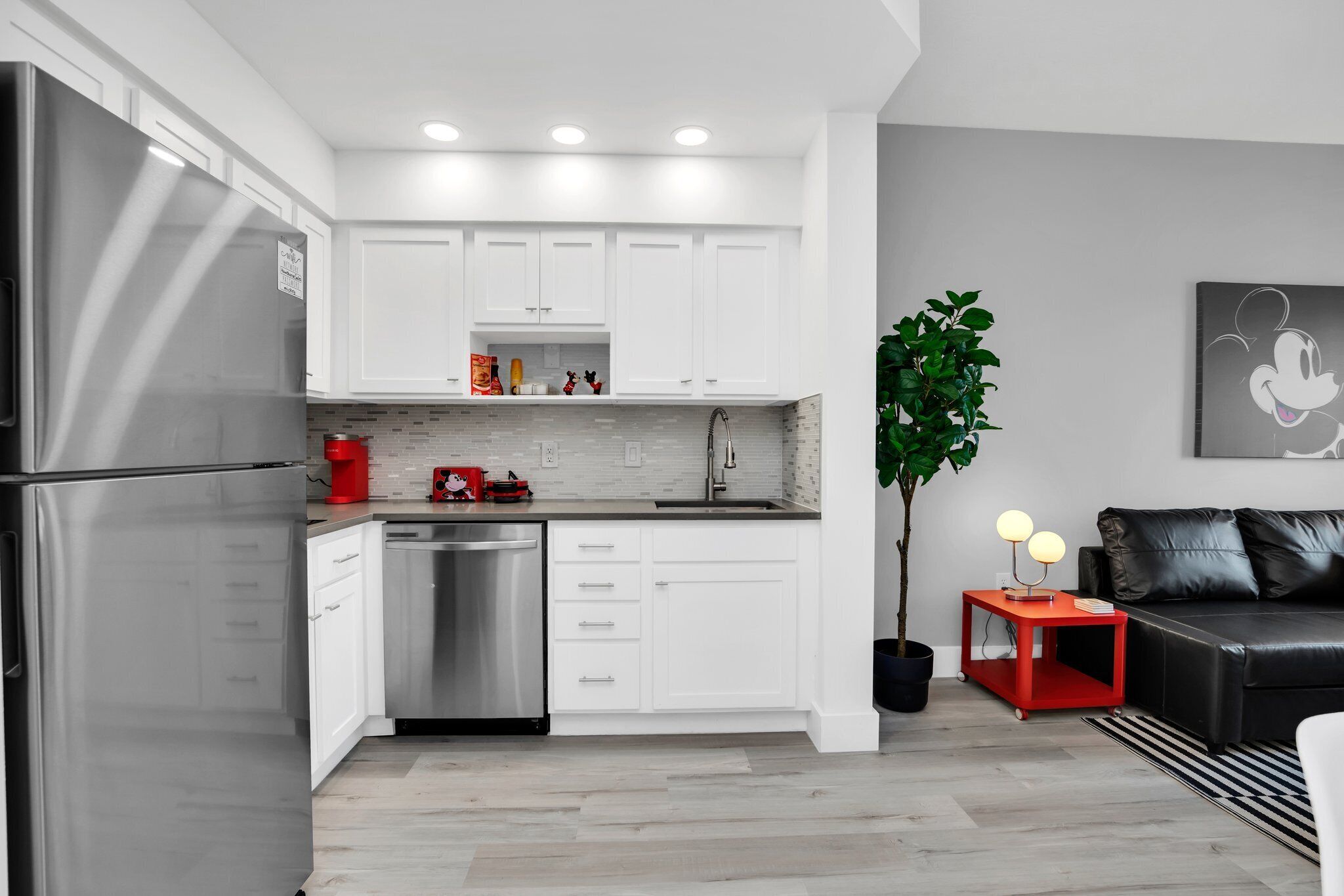 Une cuisine moderne avec des meubles blancs, des appareils en acier inoxydable et une table d’appoint rouge agrémentée de deux lampes sphériques.