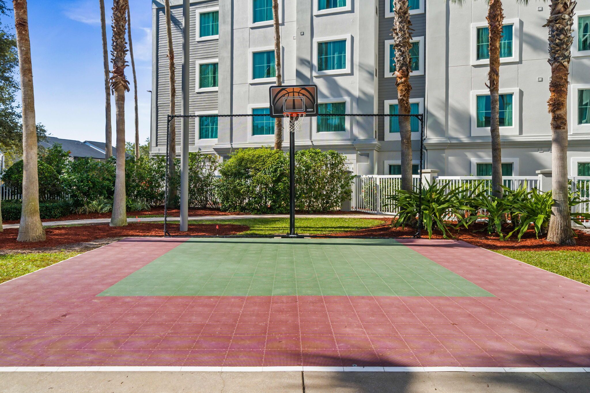 Una cancha de baloncesto con superficie verde y roja, rodeada de palmeras y un edificio con varias ventanas.