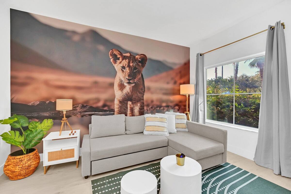 A modern living room with a large wall mural of a lion, a grey sofa, a white side table with a lamp, a green plant, and a view of greenery through a window.