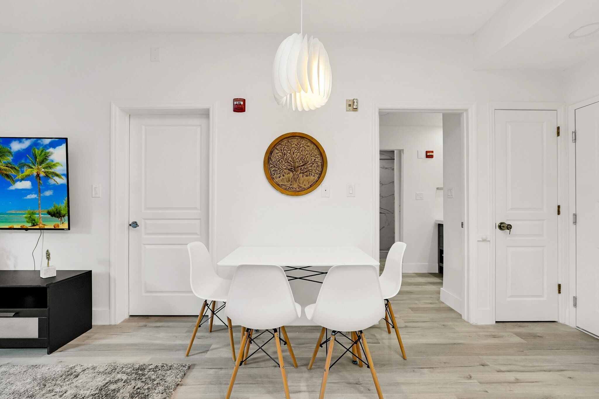 A modern dining area with a white table, four white chairs, a decorative wall piece, and a ceiling light.