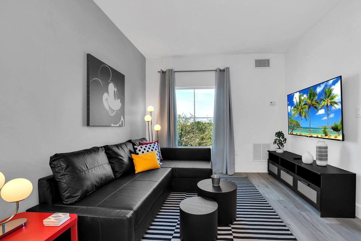 A modern living room with a black leather sofa, a red side table, a black and white striped rug, and a large flat-screen TV on the wall.