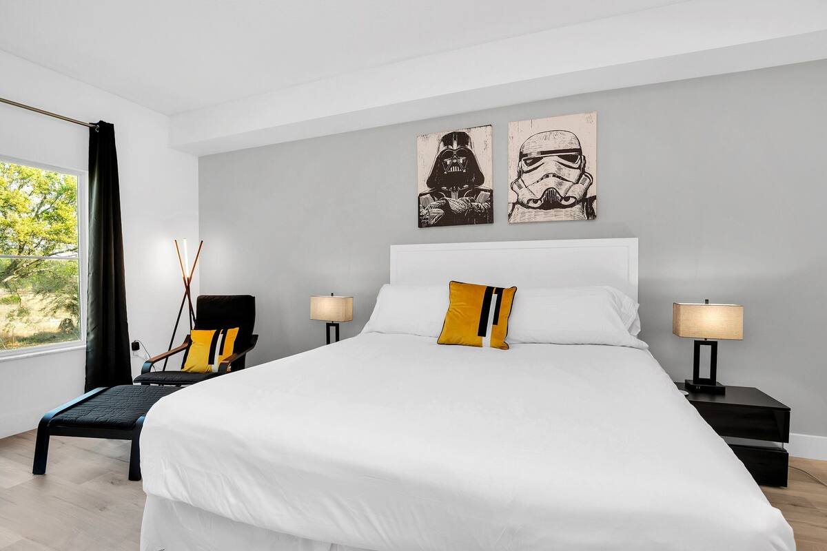 A modern bedroom with a large bed, two bedside tables with lamps, a chair, and Star Wars-themed artwork on the wall.