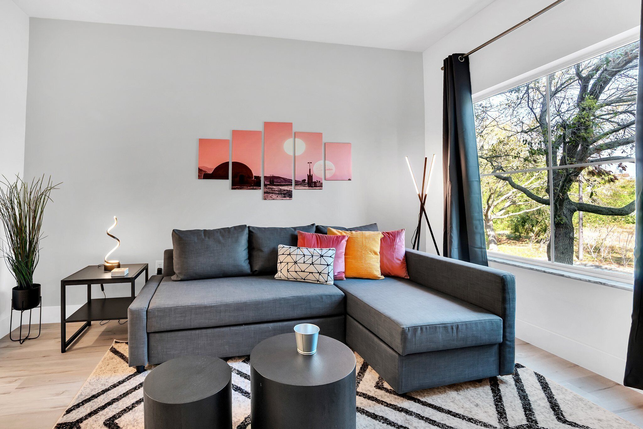 A modern living room with a grey sectional sofa, a black coffee table, a black side table with a lamp, and a large window with a view of trees.