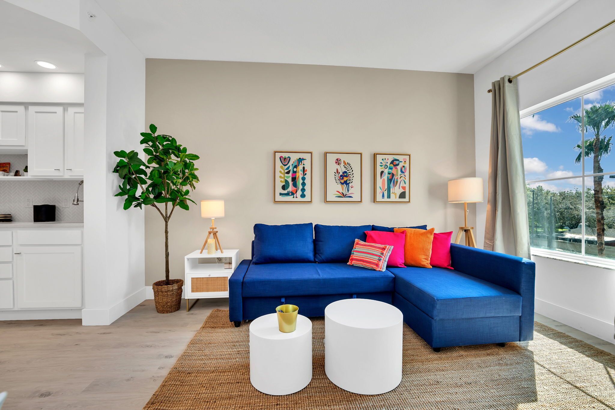 Un salon moderne avec un canapé bleu, une table basse blanche et une grande fenêtre donnant sur des palmiers.