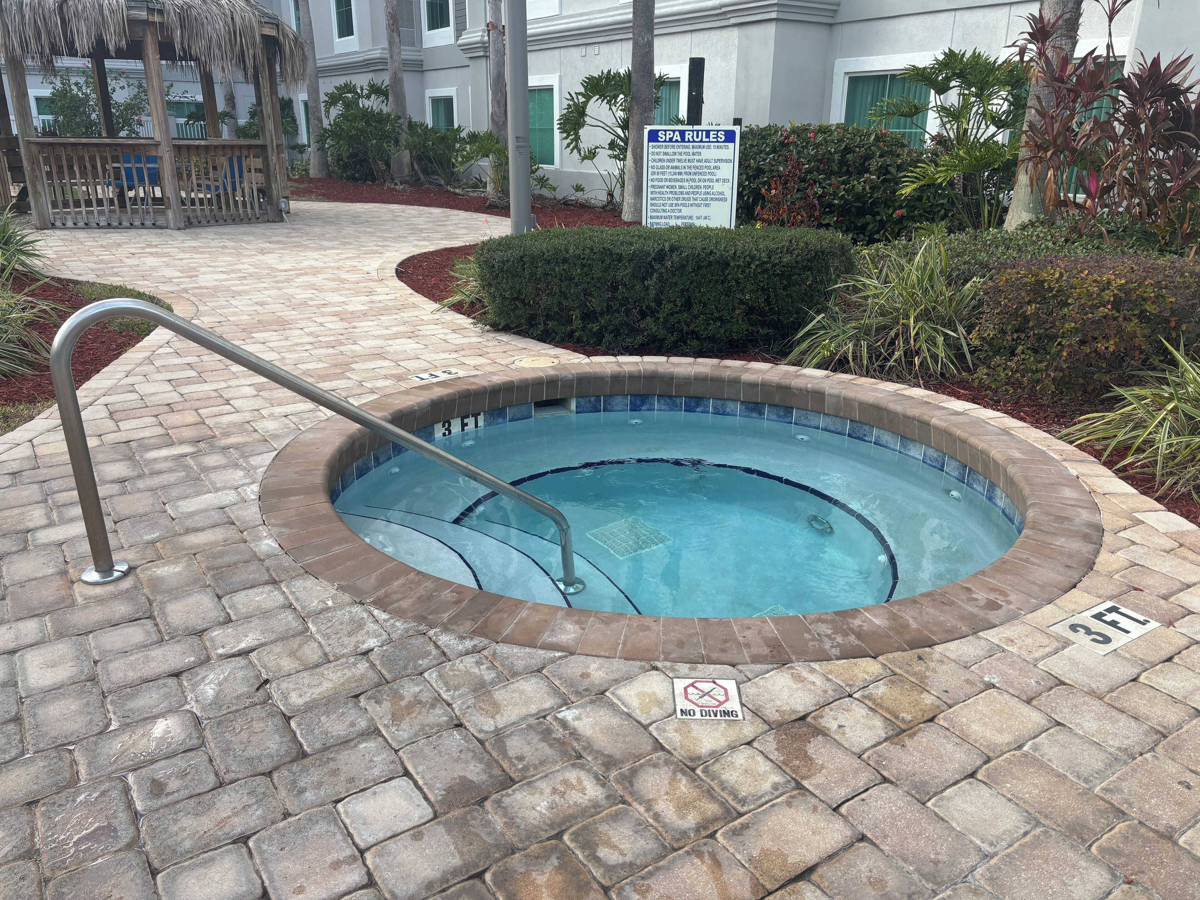 A hot tub with a 3 FT depth sign, surrounded by a brick patio and a poolside area with a wooden deck and greenery.