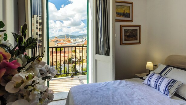 A hotel room with a bed, a nightstand, a lamp, and a view of a cityscape through a balcony. - Family run bed and breakfast. (Alghero)