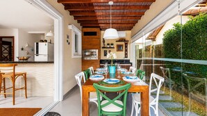 A dining area with a wooden table and chairs, a kitchen in the background, and a view of a garden through glass doors.
