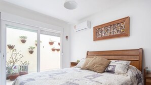 A bedroom with a bed, a wall-mounted air conditioner, and a window with potted plants.