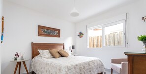 Une chambre à coucher avec un lit, une table de chevet ornée d’un vase, une fenêtre avec des rideaux et un tableau accroché au mur.