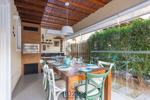 A dining area with a wooden table set for six, surrounded by white chairs. A modern kitchen with a built-in oven and a hanging light fixture is visible in the background. - Bz30 Casa em Geribá 250 metros da praia (Buzios)
