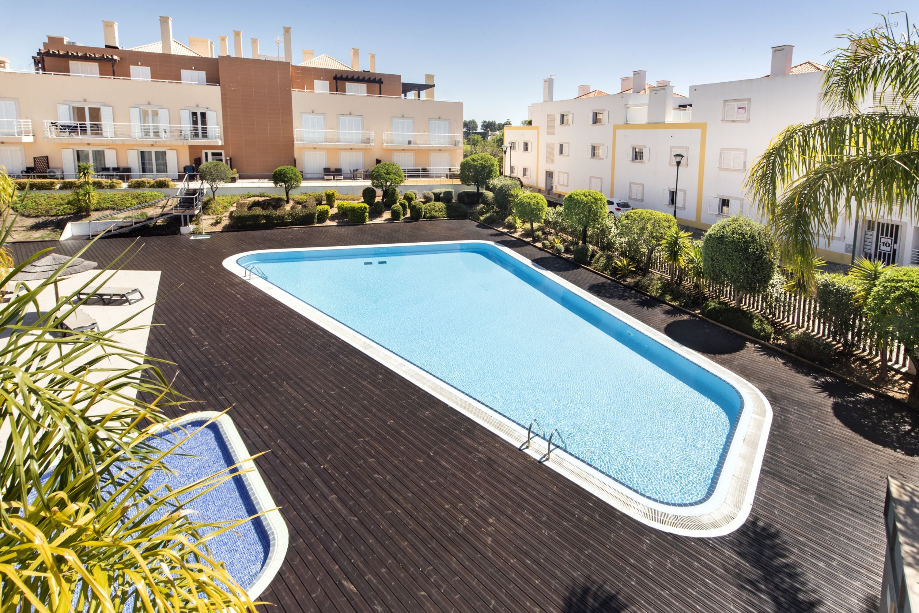 Une piscine rectangulaire entourée d’une terrasse en bois, située dans un espace aménagé avec des arbustes soigneusement taillés et quelques palmiers.