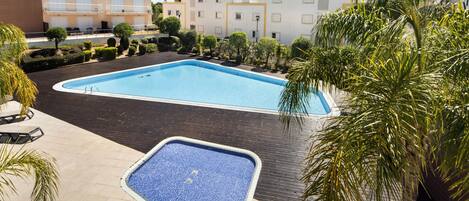 A swimming pool with a tiled deck, surrounded by neatly trimmed bushes and palm trees.