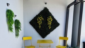A small outdoor seating area with two yellow chairs and a table, a decorative wall with green plants and a black diamond-shaped design, and a tiled floor.