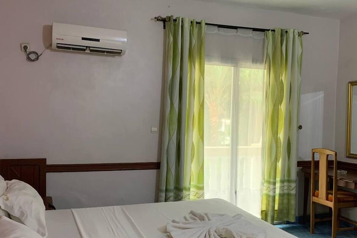 A hotel room with a bed, an air conditioning unit, a wooden chair, and a mirror.