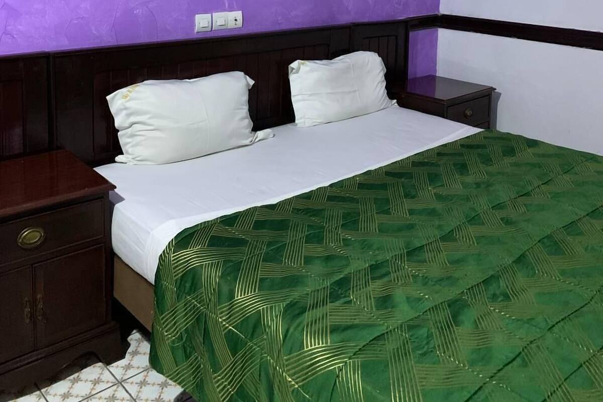 A bedroom with a purple wall, a bed with white bedding, a green patterned bedspread, a wooden nightstand, and a tiled floor.