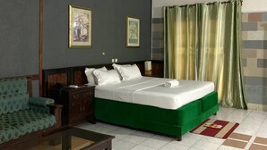 Habitación de hotel con una cama verde, una mesita de noche de madera, un sofá verde y dos cuadros en la pared.