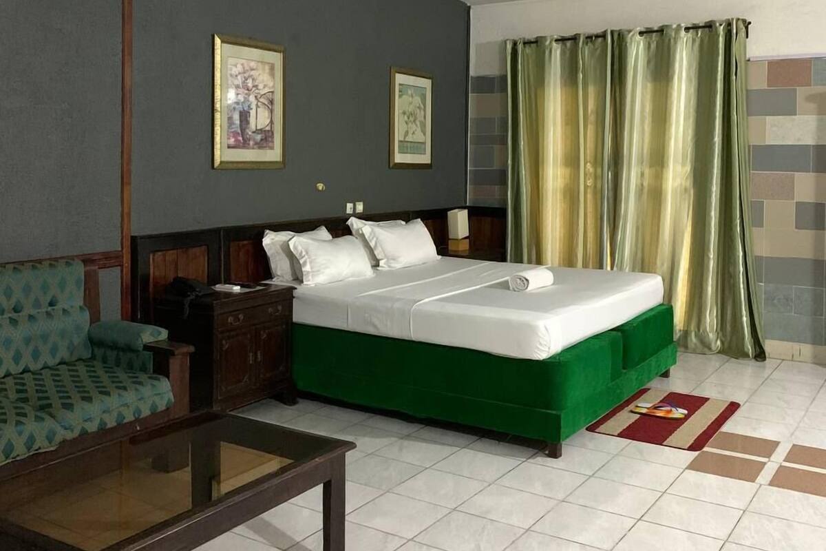 A hotel room with a green bed, a wooden nightstand, a green sofa, and two framed pictures on the wall.