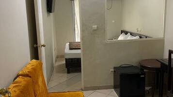 A compact room with a yellow sofa, a small table, a chair, a bed, and a mirror.