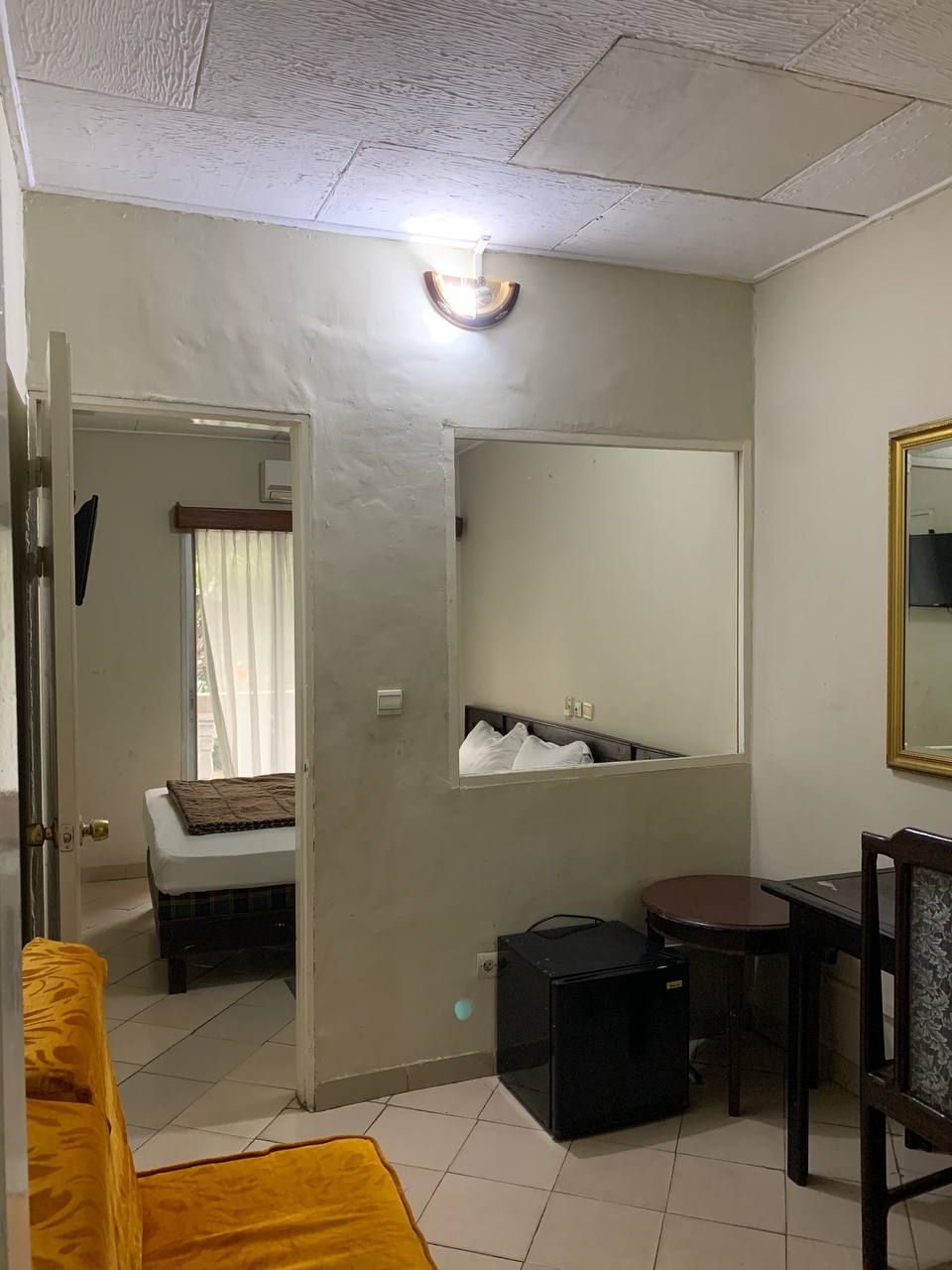 Une petite pièce comprenant un lit, une petite table, une chaise, un miroir et une fenêtre avec des rideaux.