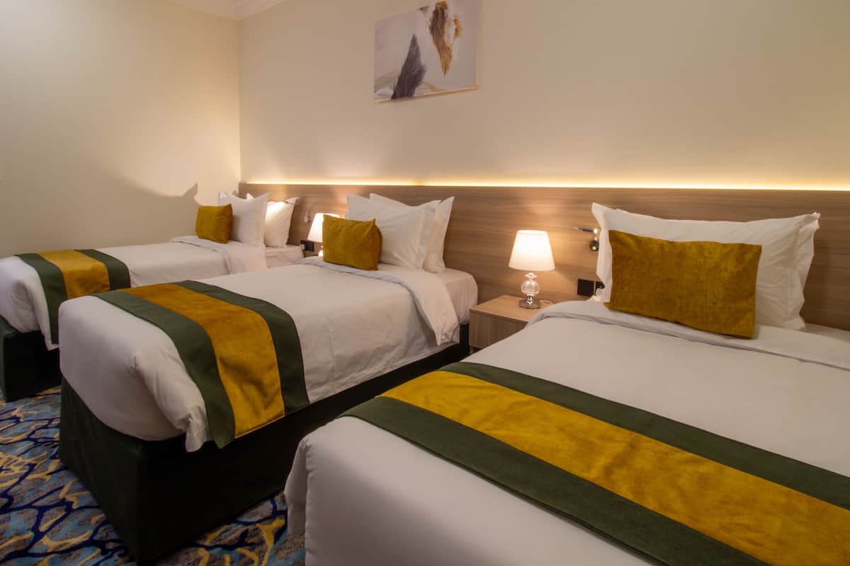 Une chambre d’hôtel avec trois lits, chacun agrémenté de draps blancs et de touches de vert et de jaune. On y trouve des lampes de chevet et un tableau au mur.