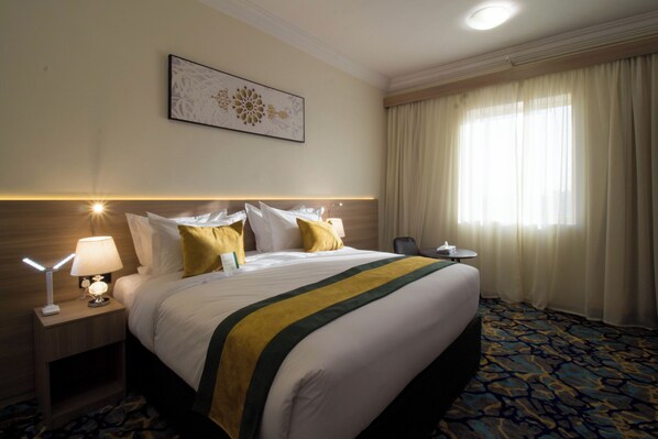 Habitación de hotel con una cama grande, una mesita de noche con lámpara, una pequeña mesa y una ventana con cortinas.