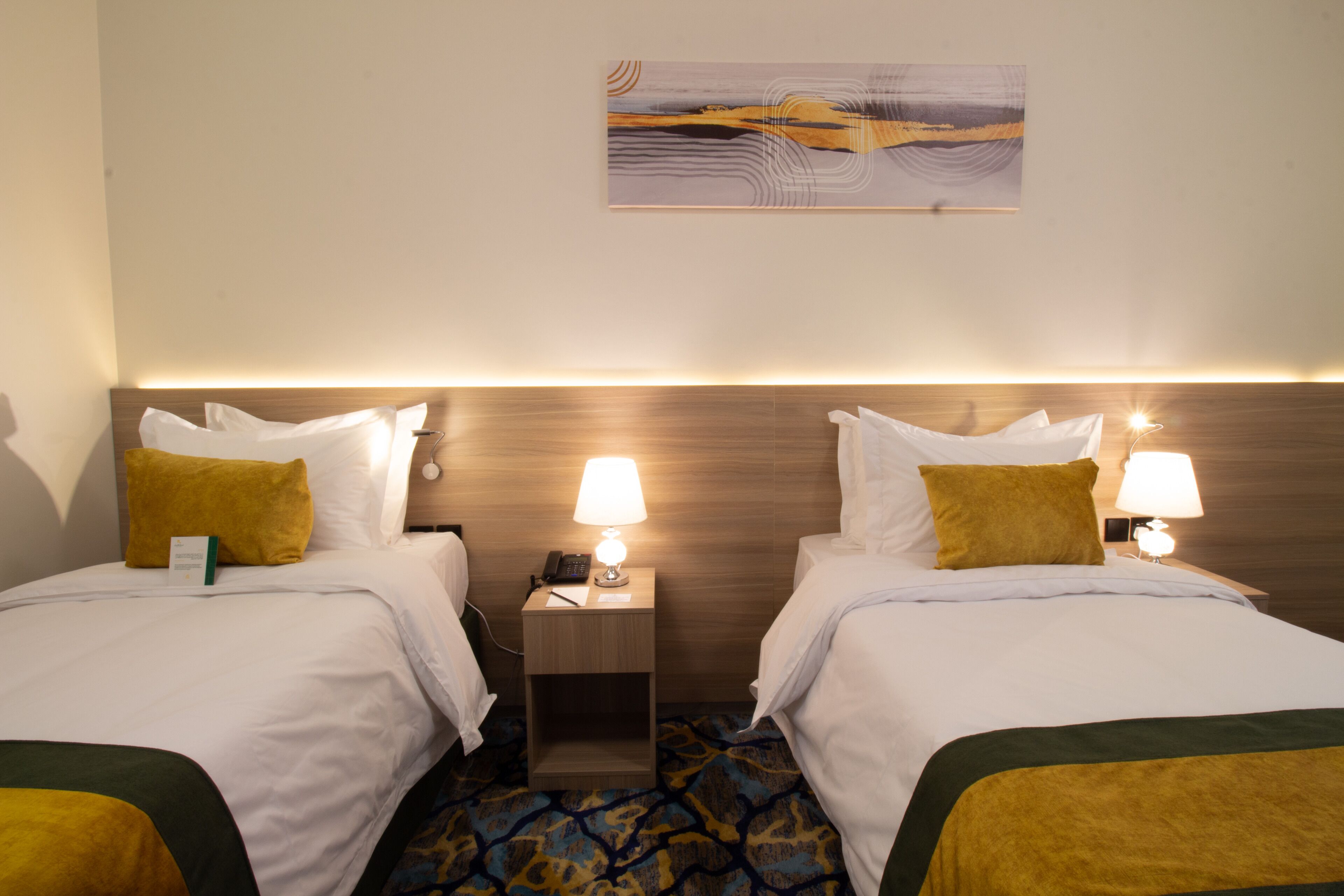 A hotel room with two beds, each with white bedding and a mustard-colored throw. There are bedside lamps, a nightstand with a phone, and a painting on the wall.