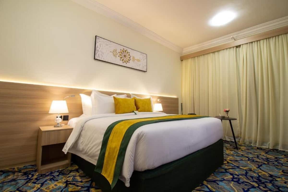 Une chambre d’hôtel avec un grand lit, deux tables de chevet avec des lampes, une petite table avec un vase et une grande fenêtre avec des rideaux.