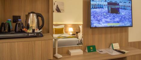 A hotel room with a wooden desk, a television displaying a cityscape, a bed with a yellow pillow, and a coffee maker.