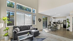 A spacious living area with a grey sectional sofa, a coffee table, and a dining table with chairs. There are large windows with blinds, a kitchen area with white cabinets, and a dining area with a view of the outdoors. - “NEW” Half Moon Retreat / +Pool Table / - 5 min away from GRATON casino (Santa Rosa)