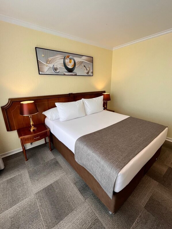 A neatly made bed with white linens and a gray throw, a wooden headboard, a bedside lamp, and a framed abstract painting on the wall. - Ocean Boulevard (Iquique)
