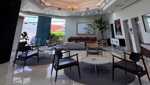 Un hall d’hôtel doté d’un plafond haut, d’un éclairage encastré, d’un lustre central, d’un espace salon avec des canapés et des fauteuils, et d’une grande fenêtre offrant une vue sur l’extérieur.