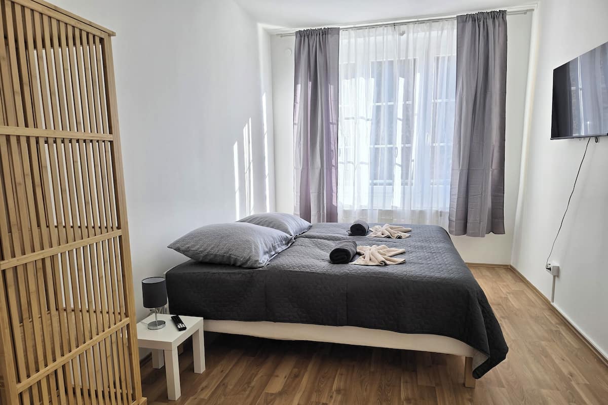 Quarto com cama, guarda-roupa de madeira, mesa de cabeceira com abajur e televisão na parede.