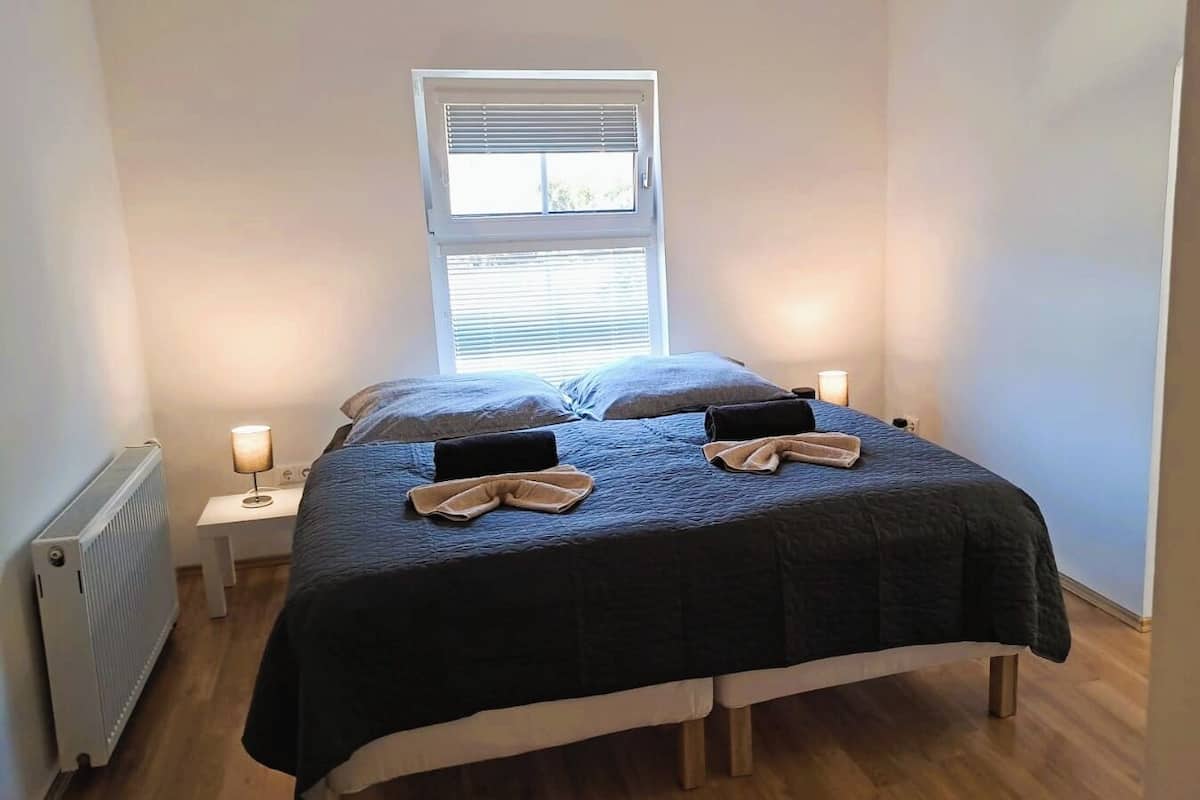 Um quarto pequeno e acolhedor com uma cama de solteiro, uma janela com persianas, uma mesa de cabeceira com abajur e um radiador.