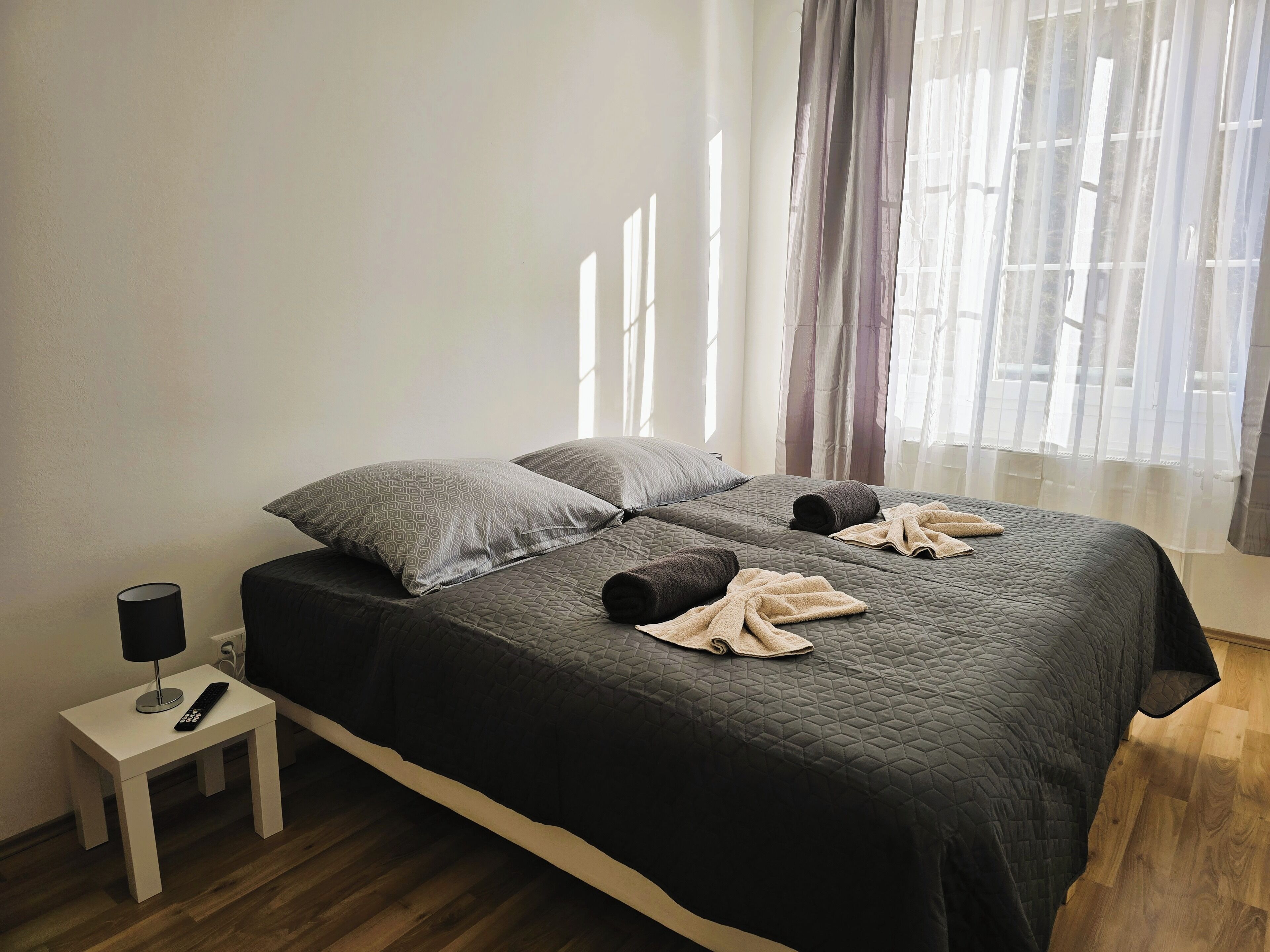 Una cama bien hecha con un edredón oscuro, dos almohadas y toallas enrolladas. Una mesita de noche blanca con una lámpara y un control remoto está al lado de la cama.