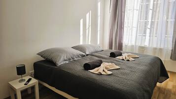 Una cama bien hecha con un edredón oscuro, dos almohadas y toallas enrolladas. Una mesita de noche blanca con una lámpara y un control remoto está al lado de la cama.