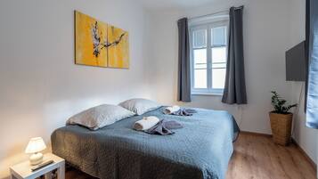 A neatly arranged bedroom with a bed, pillows, a bedside table with a lamp, a painting on the wall, a window with curtains, and a potted plant.