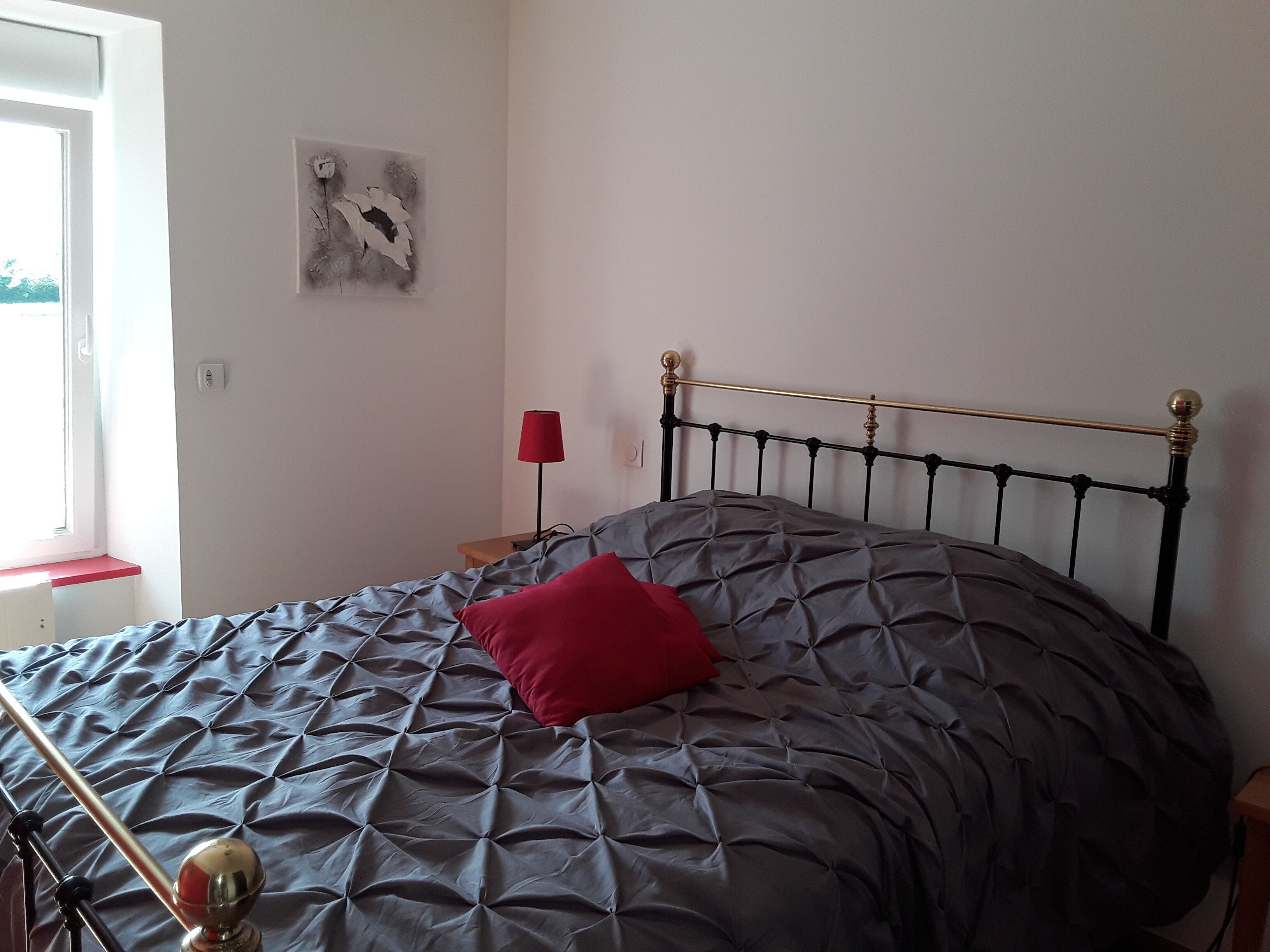 Un lit avec une couette grise et un oreiller rouge, une lampe de chevet et une œuvre d’art accrochée au mur.