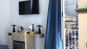 A modern hotel room with a flat-screen TV mounted on the wall, a compact kitchenette with a coffee maker, and a view of the city through a balcony with a blue curtain. - Sun&City Naples (Naples)