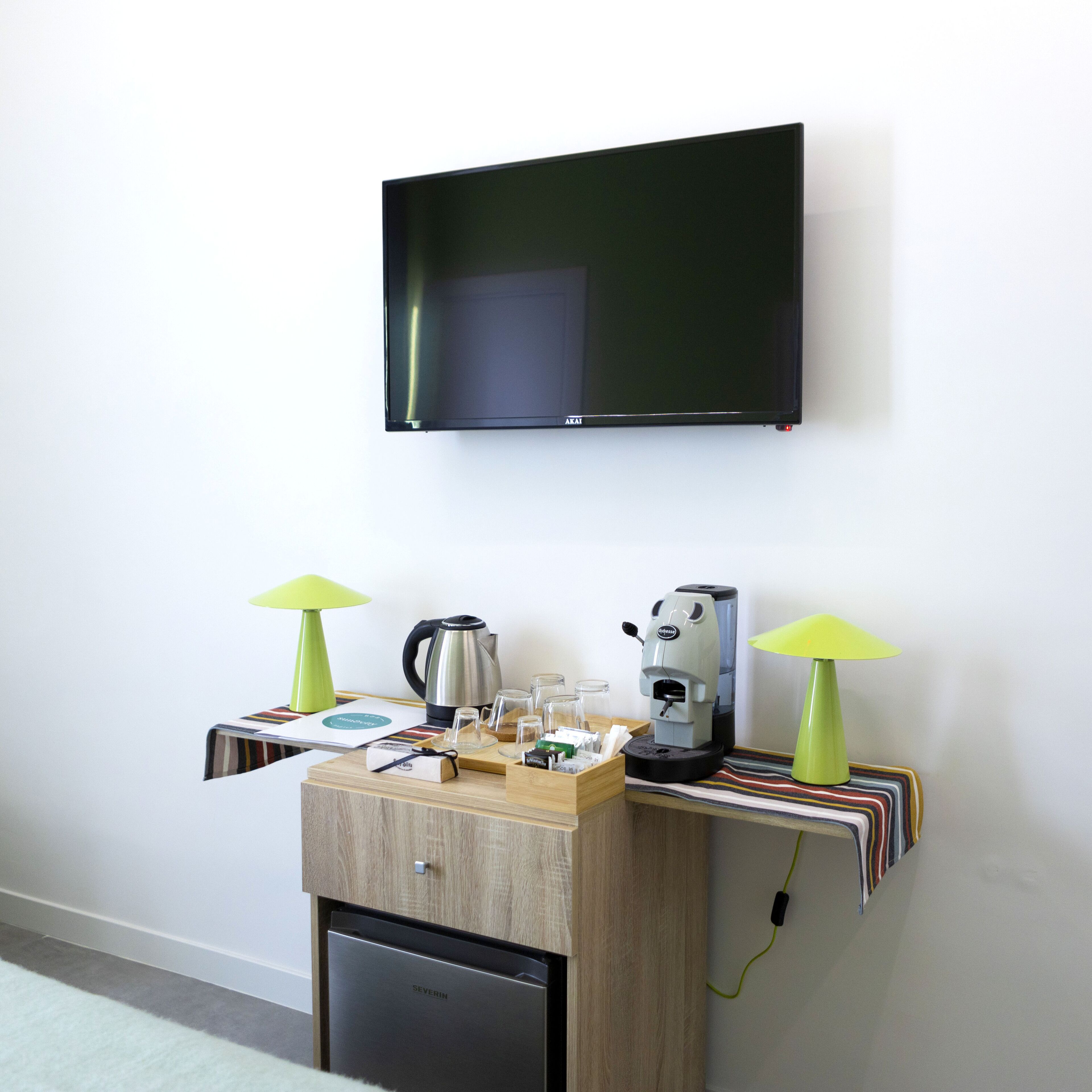 A room with a flat-screen TV mounted on the wall, a coffee machine, a kettle, and two lamps on a wooden table.