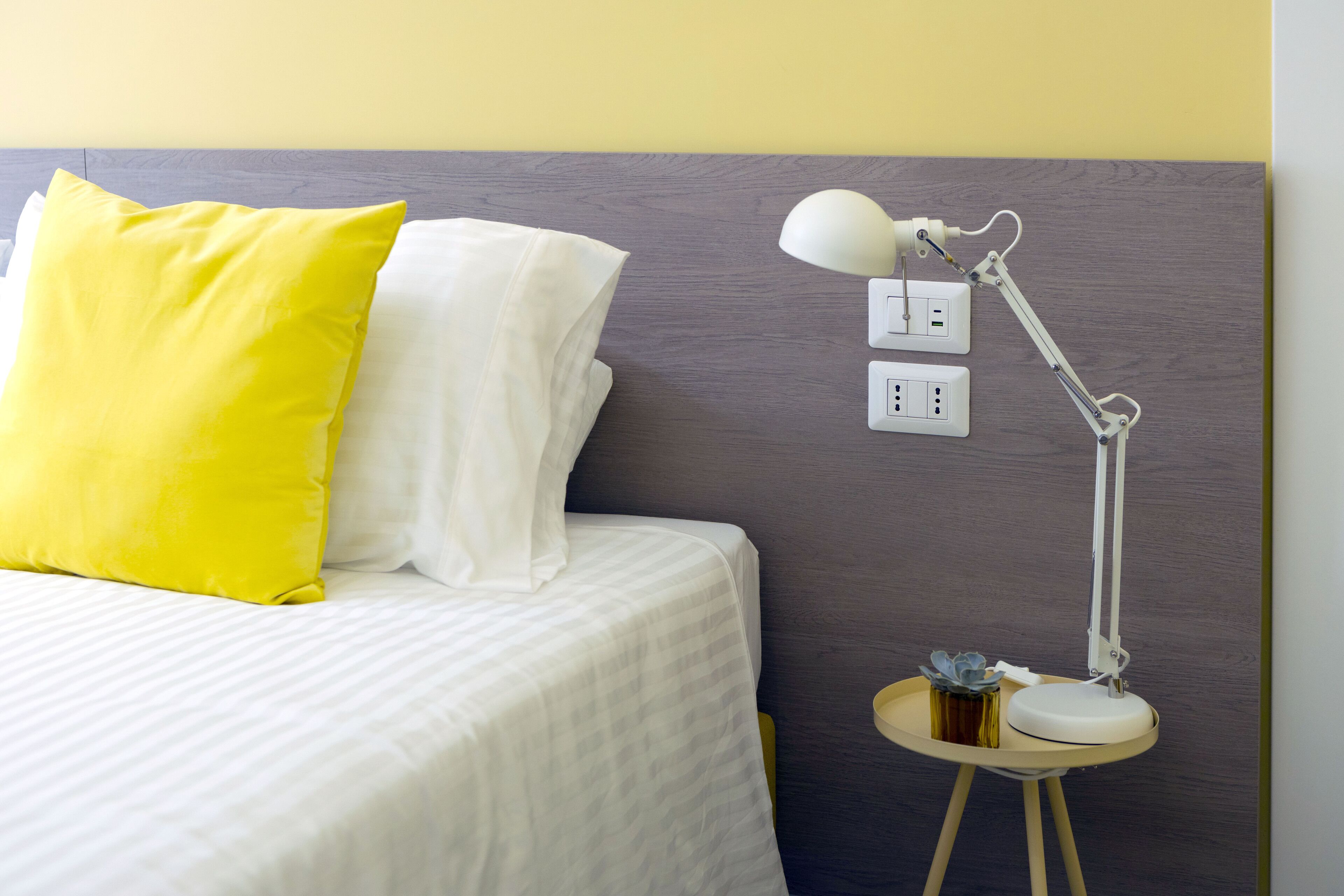 A neatly made bed with a white and yellow pillow, a bedside table with a lamp, and a power outlet on the wall.