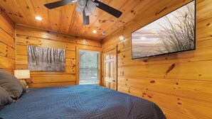 A wooden cabin room with a bed, a ceiling fan, a painting, and a window. - Couples Refuge by Ghosal Luxury Lodging (Sevierville)
