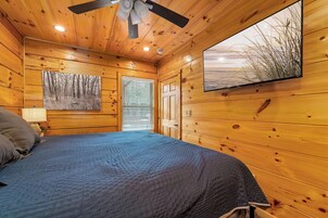 A wooden cabin room with a bed, a ceiling fan, a painting, and a window.