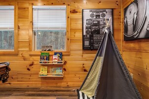 A cozy cabin interior with wooden walls, a bookshelf with children's books, a hammock, and a wall-mounted radio.