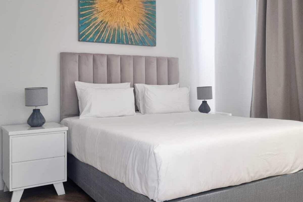 Une chambre à coucher moderne avec un lit recouvert d’un tissu gris, une literie blanche et un grand tableau abstrait accroché au mur.